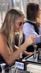 Smiling woman in sunglasses stirring a cocktail at an urban rooftop bar, wearing gold beaded bracelets; jigger and mixing tools on a tray with a city skyline blurred in the background.