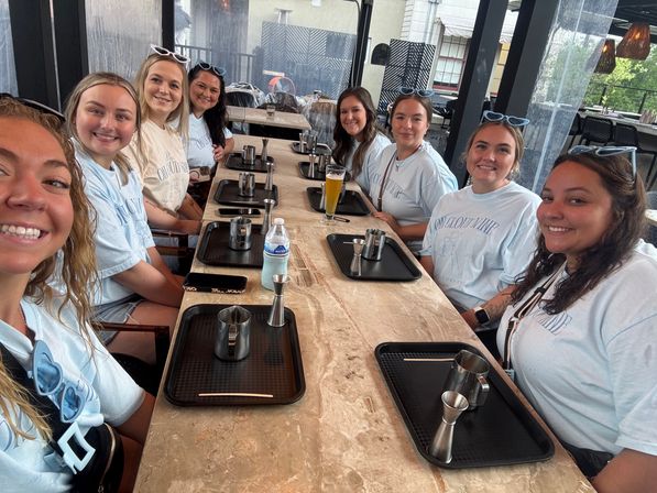 Eight friends in matching light blue shirts smiling for a selfie around a long marble table on a covered outdoor restaurant patio, each with black service trays, metal jiggers and cups, a water bottle and a beer glass—casual brunch gathering.