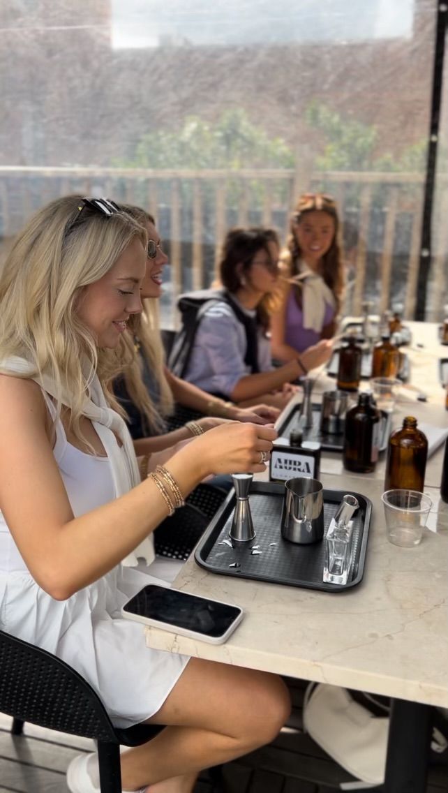 Group of friends at a sunny rooftop patio cocktail-making class, seated at a marble bar with jiggers, stainless steel pitchers, amber bottles and trays