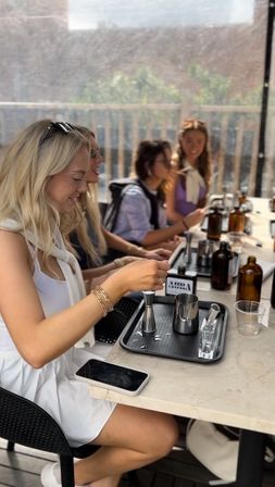Group of friends at a sunny rooftop patio cocktail-making class, seated at a marble bar with jiggers, stainless steel pitchers, amber bottles and trays