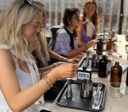 Group of friends at a sunny rooftop patio cocktail-making class, seated at a marble bar with jiggers, stainless steel pitchers, amber bottles and trays