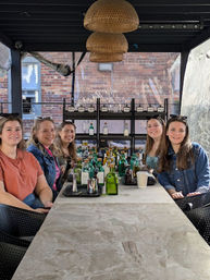 Five friends seated at a long stone table on an urban covered patio with woven pendant lights, surrounded by bottles and trays for a daytime spirits tasting against a brick-wall backdrop.