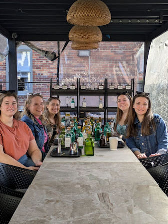 Five friends seated at a long stone table on an urban covered patio with woven pendant lights, surrounded by bottles and trays for a daytime spirits tasting against a brick-wall backdrop.
