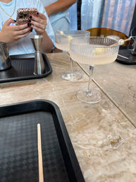 Two pale cocktails in ribbed coupe glasses with pearl-topped skewers on a marble table, with serving trays and hands holding a leopard-print phone in the background.