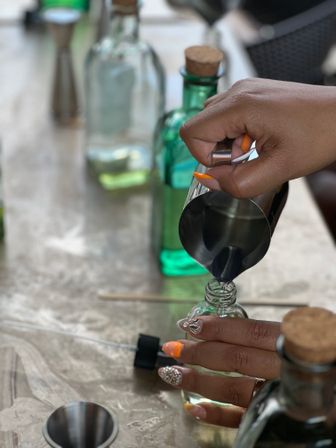Close-up of hands with bright orange and glittery nails pouring liquid from a stainless steel jigger into a small glass bottle, surrounded by green corked bottles and bar tools on a tabletop — cocktail/mixology scene.