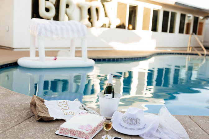 Sunlit pool deck bachelorette setup with a rhinestone "BRIDE" hat and veil, pink-sprinkled cake, glass of champagne and bottle in an ice bucket, plus a striped inflatable float in the pool
