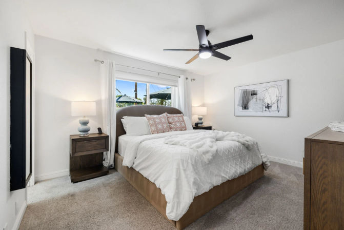 Bright modern coastal bedroom with upholstered queen bed, white textured bedding, wooden nightstands with lamps, black ceiling fan, and a window showing palm trees and blue sky.