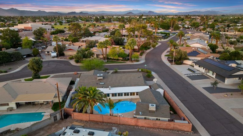 Aerial sunset of a desert suburb: single-story homes on curving streets, a bright blue backyard pool with palm trees, xeriscaped yards, and distant mountains on the horizon.