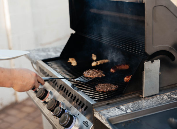 Hand flipping sizzling burger patties and sausages on a stainless-steel gas grill with smoke rising on a backyard patio