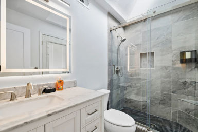 Bright modern bathroom with marble vanity and LED-framed mirror, white toilet, polished chrome faucets, and glass-enclosed walk-in shower with gray stone tiles and handheld rain showerhead.