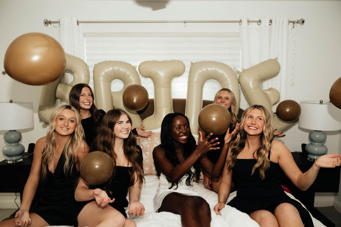 Bachelorette party in a cozy bedroom: bride in white and bridesmaids in black sitting on a bed, tossing gold balloons with beige letter balloons spelling BRIDE behind them.