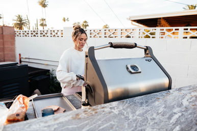 Young woman in a white sweatshirt grilling burger patties on a stainless-steel gas grill in a sunny backyard patio with palm trees and a white cinderblock wall