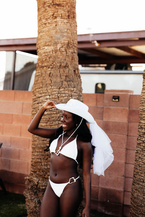 Smiling woman in a white bikini and white cowboy hat with attached bridal veil, adjusting her hat beside a palm tree and brick wall on a sunny outdoor patio.