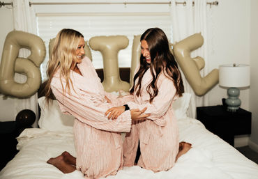 Two women kneeling on a bed in matching pink plush robes, laughing and holding hands with beige party balloons and white bedroom decor in the background