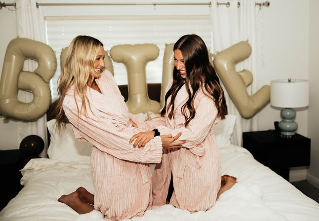 Two women kneeling on a bed in matching pink plush robes, laughing and holding hands with beige party balloons and white bedroom decor in the background