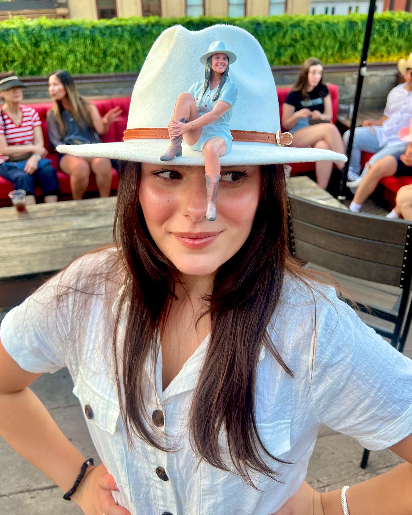 Playful rooftop patio scene: woman in a white wide-brim hat with a tiny doll-like figure perched on the brim, wearing a white button shirt and smiling, with friends seated on red benches blurred in the background.