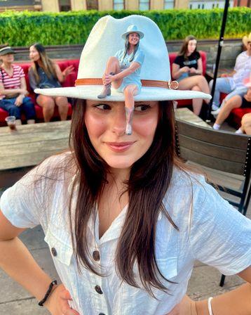 Playful rooftop patio scene: woman in a white wide-brim hat with a tiny doll-like figure perched on the brim, wearing a white button shirt and smiling, with friends seated on red benches blurred in the background.