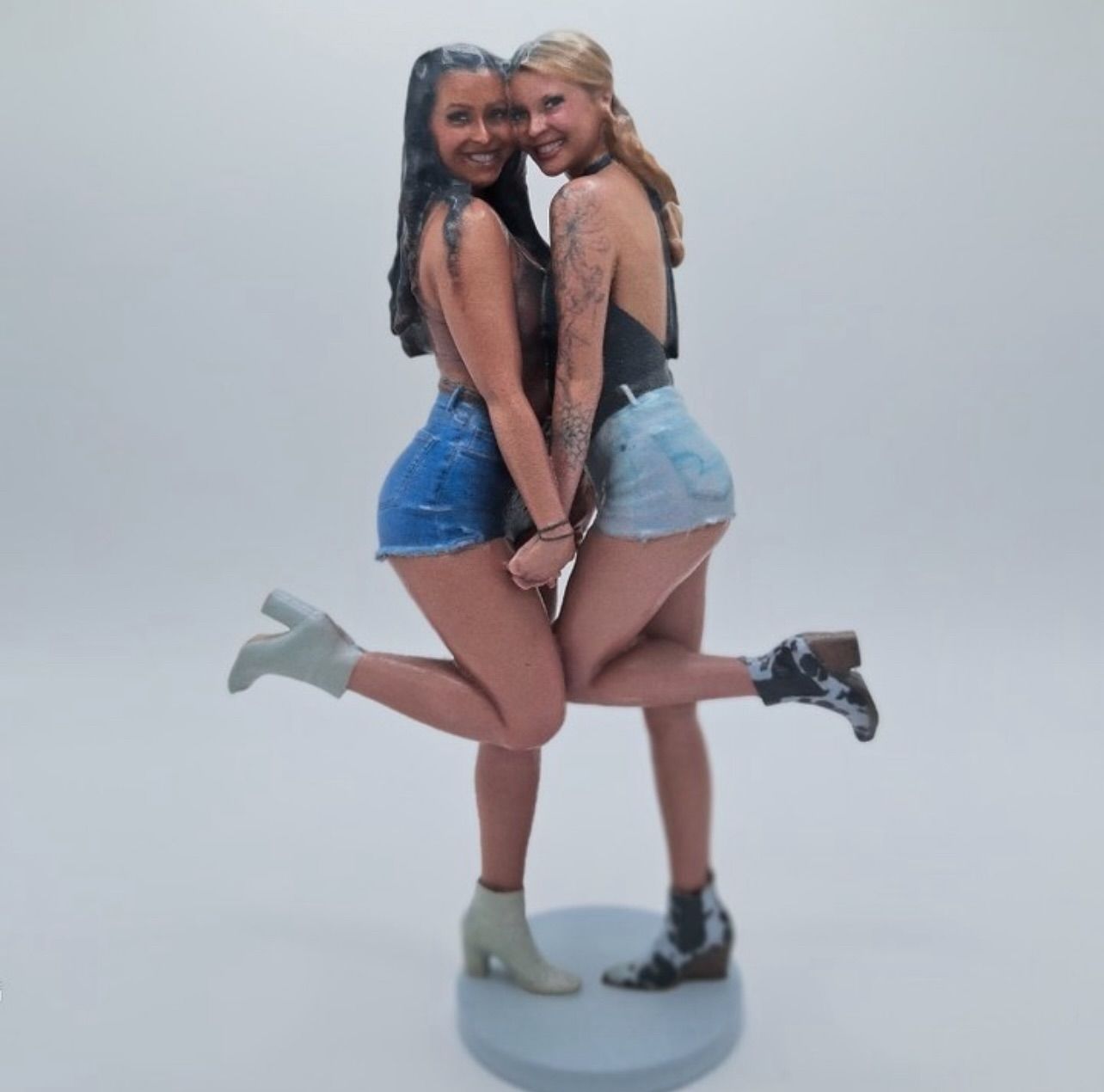Playful studio portrait of two friends posing back-to-back with linked hands and one leg lifted, wearing sleeveless tops, denim shorts and ankle boots against a plain light background.