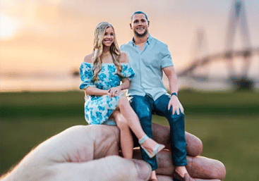 Hand holding a miniature couple figurine seated together at sunset in a park with a blurred bridge in the background; woman in a blue floral dress and man in a light shirt and jeans.