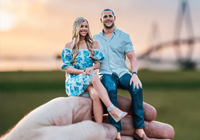 Hand holding a miniature couple figurine seated together at sunset in a park with a blurred bridge in the background; woman in a blue floral dress and man in a light shirt and jeans.