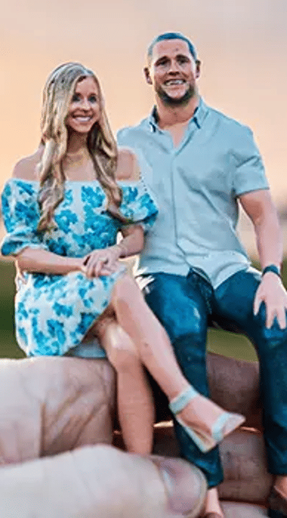 Hand holding a miniature couple figurine seated together at sunset in a park with a blurred bridge in the background; woman in a blue floral dress and man in a light shirt and jeans.
