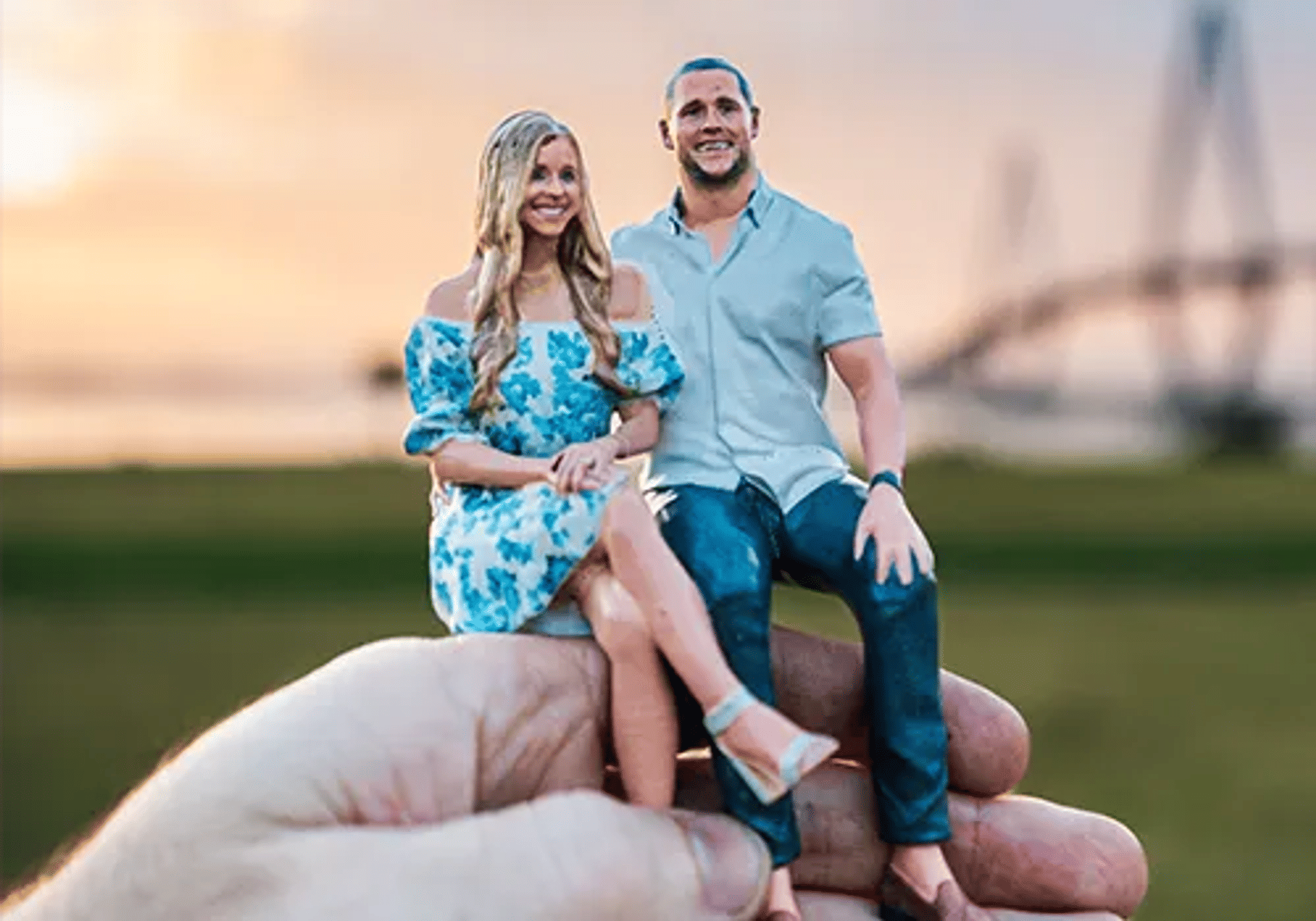 Hand holding a miniature couple figurine seated together at sunset in a park with a blurred bridge in the background; woman in a blue floral dress and man in a light shirt and jeans.