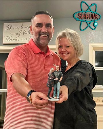 Smiling couple at home holding a tiny custom 3D-printed figurine of themselves, man in a coral polo and woman in a dark top standing in a cozy living room.