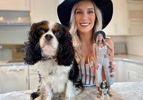 Smiling woman with long blonde hair and black wide‑brim hat in a bright kitchen holding a striped‑jumpsuit miniature figurine, with a tricolor Cavalier King Charles Spaniel sitting on the countertop