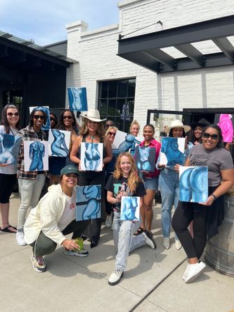 Cheerful outdoor painting workshop on a sunny urban patio in front of a white‑brick building, a group of adults proudly holding blue‑toned figurative torso canvases.