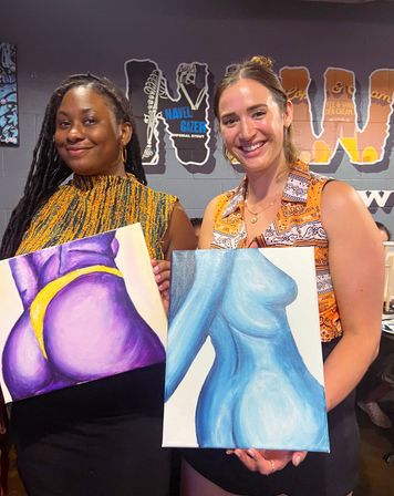 Two smiling women at a paint-night event holding colorful canvas paintings of abstract torsos—one purple with a yellow accent and one blue—against a casual studio backdrop.
