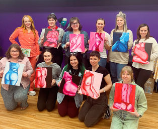 Group of eleven adults in a colorful art studio holding vibrant figurative paintings of female torsos in pinks and blues, posing against a purple wall and wood floor.