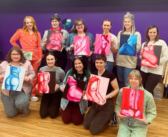 Group of eleven adults in a colorful art studio holding vibrant figurative paintings of female torsos in pinks and blues, posing against a purple wall and wood floor.
