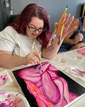 Smiling artist with glasses painting a vibrant pink abstract floral on canvas in an indoor art studio, holding multiple paintbrushes during a paint-night workshop.