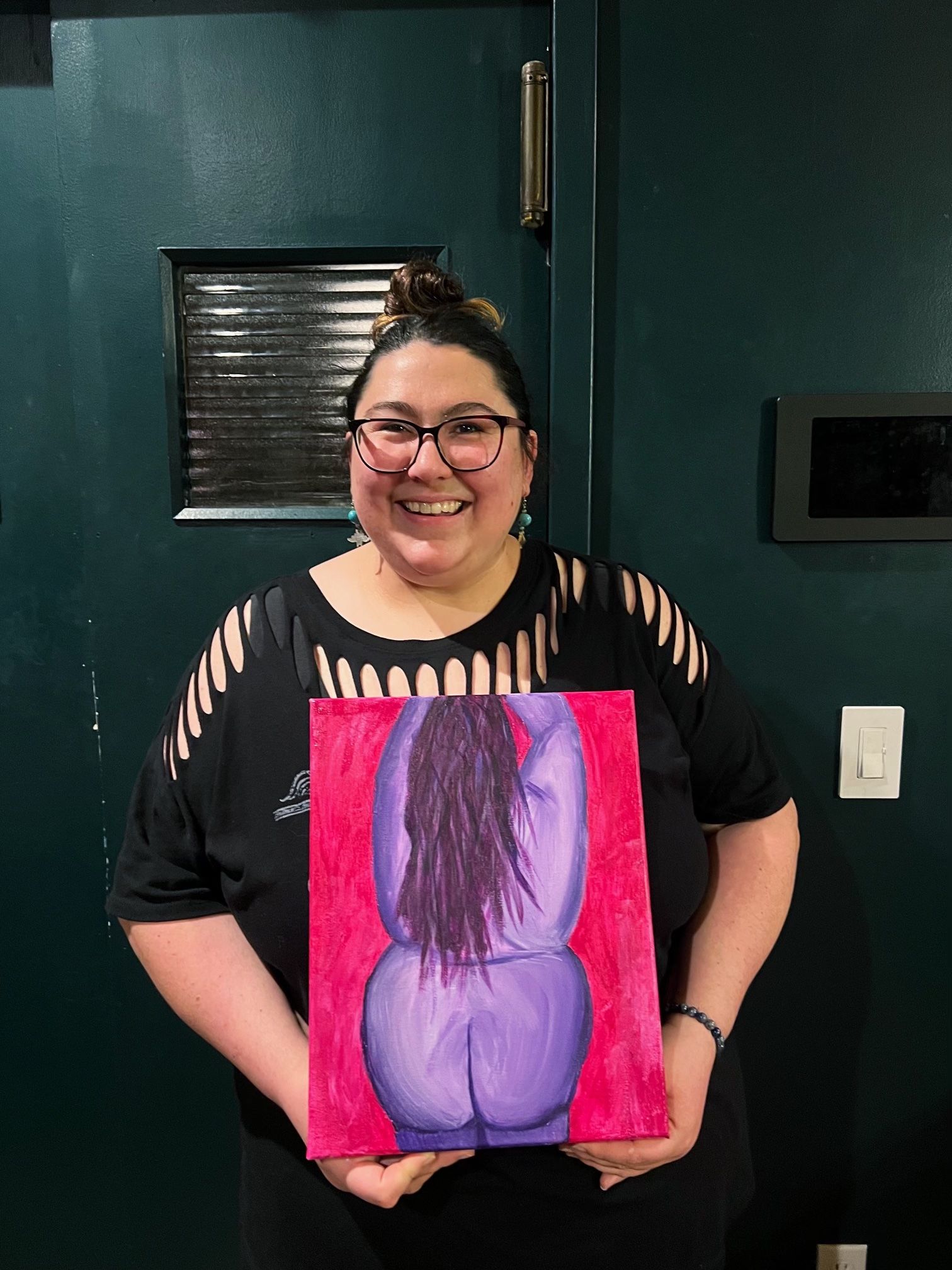 Smiling person with glasses and a topknot holding a colorful canvas painting of a stylized purple figure’s back on a red background, wearing a black cutout-sleeve shirt and standing indoors against a dark green door.