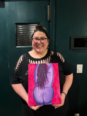 Smiling person with glasses and a topknot holding a colorful canvas painting of a stylized purple figure’s back on a red background, wearing a black cutout-sleeve shirt and standing indoors against a dark green door.