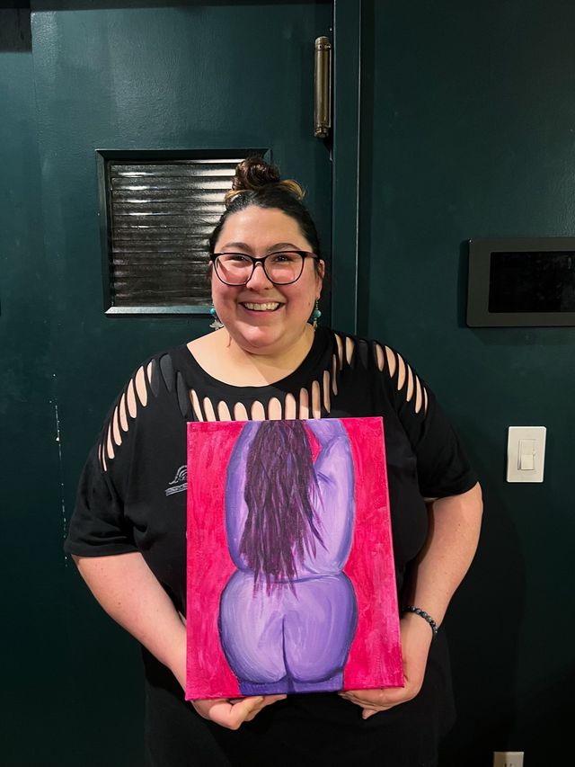 Smiling person with glasses and a topknot holding a colorful canvas painting of a stylized purple figure’s back on a red background, wearing a black cutout-sleeve shirt and standing indoors against a dark green door.