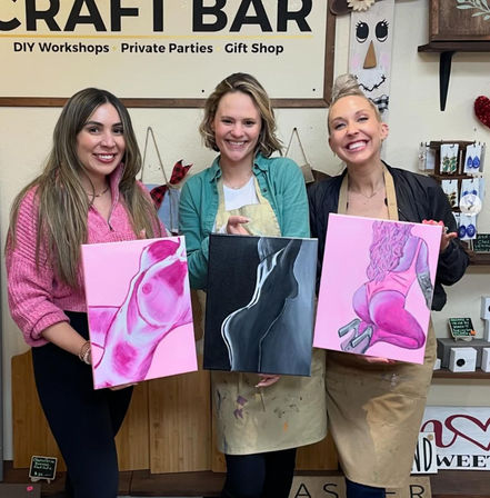 Three smiling women in aprons pose in a craft studio holding pink- and black-themed figure paintings from a paint-night DIY workshop, group painting class vibe.