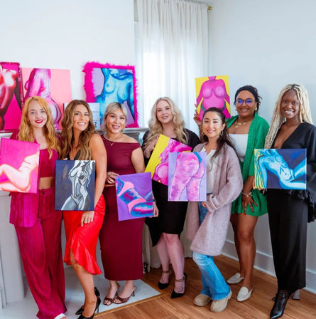 Seven women smiling in a bright home art studio holding vibrant, colorful abstract nude figure paintings against white walls and a wooden floor