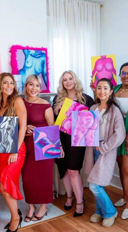 Seven women smiling in a bright home art studio holding vibrant, colorful abstract nude figure paintings against white walls and a wooden floor