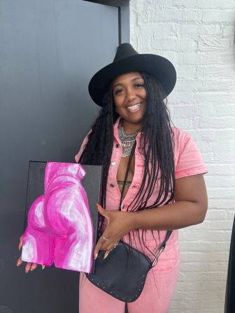 Smiling person in a black wide-brim hat and pink outfit holding a small canvas painted with a bright pink abstract torso, posed against a gray door and white brick wall.