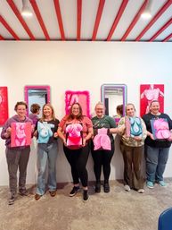 Six people in an art studio painting class holding vibrant pink and blue figurative torso paintings in front of a white gallery wall with mirrors and red ceiling beams.