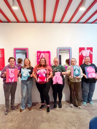 Six people in an art studio painting class holding vibrant pink and blue figurative torso paintings in front of a white gallery wall with mirrors and red ceiling beams.