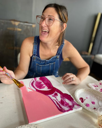 Laughing woman in denim overalls and clear glasses painting a pink and magenta acrylic canvas at an indoor art studio table with a paper-plate palette and brush.