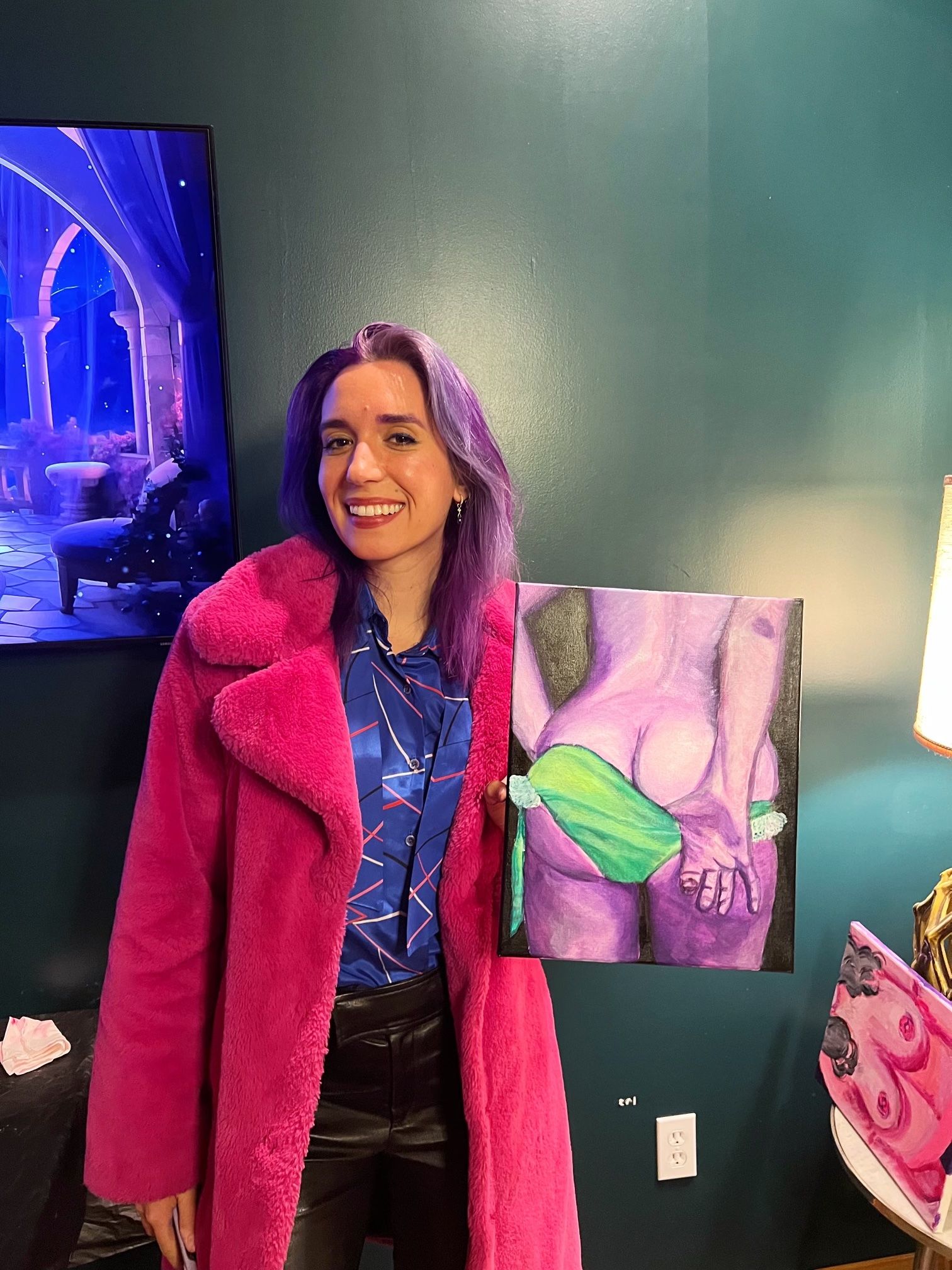 Smiling person with purple hair wearing a bright pink coat in an art studio, holding a colorful painting of a purple-toned figure tugging green underwear against a teal wall.