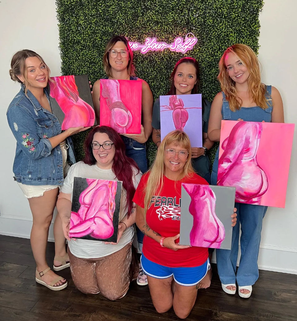 Six women at a paint-night art studio posing with pink stylized figure paintings in front of a green faux hedge wall and neon Love Yourself sign, hardwood floor visible