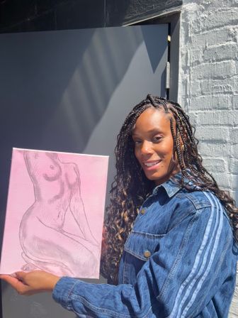 Smiling person with braided hair in a denim jacket holding a pink canvas with a pencil sketch of a seated female nude, posed by a sunlit gray door and white brick wall.