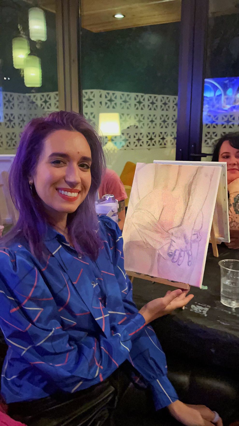 Smiling woman with purple hair at a paint-night workshop holding a canvas sketch of clasped hands in a cozy indoor evening setting.