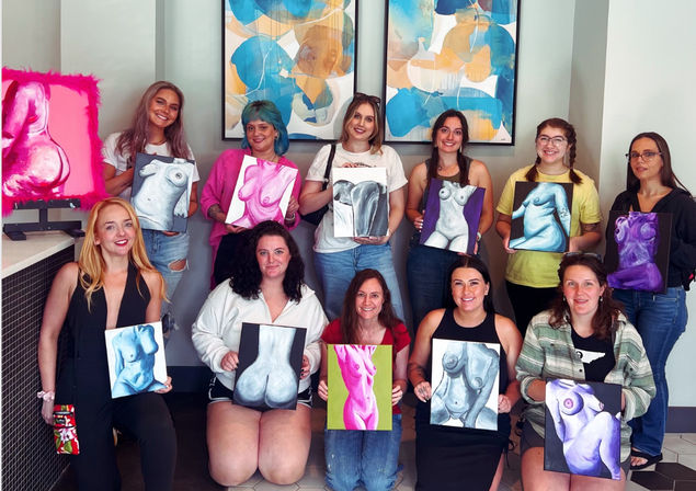 Group of women at a painting workshop in an indoor art studio, smiling and holding colorful canvases of female torsos and nudes with abstract wall art behind them.