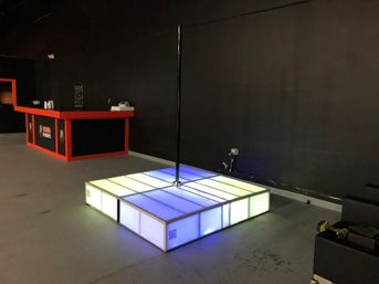 Glowing LED square dance platform with a center pole, lit in blue and white, set on a concrete floor in a dark indoor event space next to a red-trimmed bar counter.
