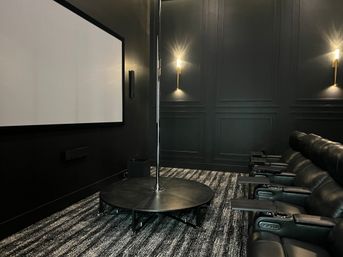 Dark-paneled home theater with blank projection screen, row of black leather reclining seats with cupholders, round stage with chrome dance pole, striped carpet and warm wall sconces.